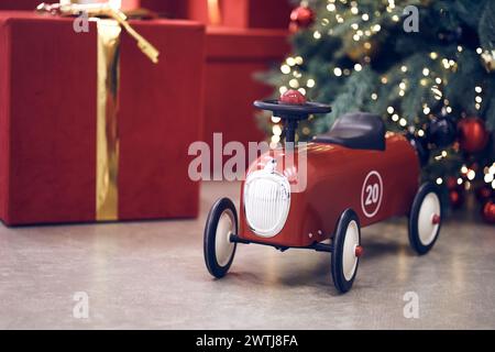 Seitenansicht des roten Retro-Autos für Kinder auf dem Hintergrund von Geschenken in roten Kästen mit goldenen Bögen, Weihnachtsbaumgirlande, Lichterketten, roten und blauen Kugeln. Hintergrund, Kopierraum, Bokeh. Stockfoto