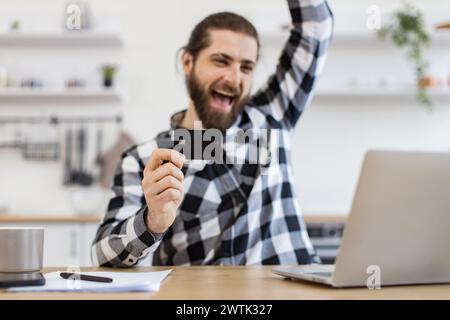 Emotionaler Erwachsener mit Debitkarte, die die Faust hebt, während er glücklich auf den Computer blickt. Stockfoto