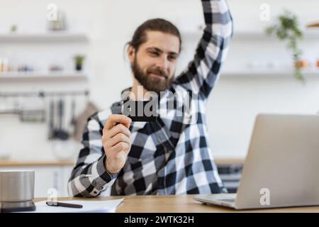 Emotionaler Erwachsener mit Debitkarte, die die Faust hebt, während er glücklich auf den Computer blickt. Stockfoto