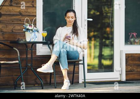 Entspannte Frau, die auf der Terrasse einer hölzernen A-Frame-Kabine mit Smartphone sitzt Stockfoto