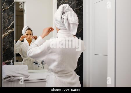 Die Frau wickelte ihr Haar nach der Dusche in ein Handtuch und sah in den Spiegel und reparierte ein Handtuch Stockfoto