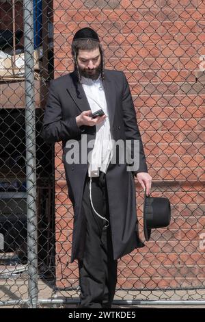 Während er auf einen Bus wartet, liest ein orthodoxer Mann von seinem Handy. Es scheint ein iPhone zu sein. In Brooklyn, New York. Stockfoto