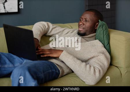 Porträt eines afroamerikanischen Erwachsenen, der einen Laptop benutzt, während er sich zu Hause oder im Büro auf der Couch entspannt Stockfoto