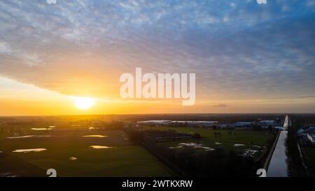 Dieses Bild bietet eine ruhige Sonnenuntergangsszene, bei der die Sonne in Richtung Horizont eintaucht und ein goldenes Licht ausstrahlt, das sich vom Wasser in den überfluteten Feldern reflektiert. Die ländliche Straße verläuft parallel zu einem Kanal und führt den Blick durch die Landschaft. Der Himmel ist mit einem Wolkenmuster geschmückt, das das Sonnenlicht streut und eine dramatische Kulisse schafft. Dieses Bild verkörpert einen friedlichen, aber kraftvollen Moment in einer ländlichen Umgebung und unterstreicht die Schönheit der Elemente der Natur. Sonnenuntergang über einer Landstraße und überfluteten Feldern. Hochwertige Fotos Stockfoto