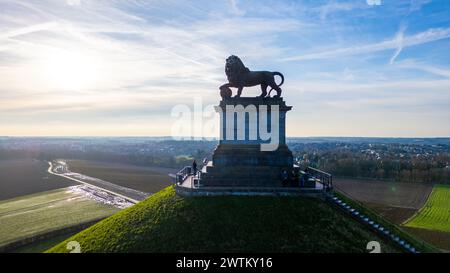 Waterloo, Brüssel, Belgien, 25. Februar 2024 das Foto zeigt den Löwenhügel mit seiner imposanten Löwenstatue an der historischen Stätte des Waterloo Schlachtfelds. Die Sonne am frühen Morgen oder am späten Nachmittag bildet eine strahlende Kulisse, die ein zartes Leuchten auf das Denkmal wirft. Der Hügel selbst ist von lebhaftem grünem Gras bedeckt, und die umliegende Landschaft ist in der Ferne sichtbar, mit Straßen und Feldern von Ackerland. Das Denkmal steht als Wächter über dem Gebiet und erinnert an die Soldaten, die in der Schlacht gekämpft haben. Besucher können auf der Treppe und dem Bahnsteig gesehen werden, was einen Eindruck von vermittelt Stockfoto