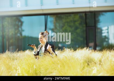 Glückliche, moderne Angestellte in der Nähe des Business Centers in schwarzer Jacke mit Smartphone und einer Tasse Kaffee. Stockfoto