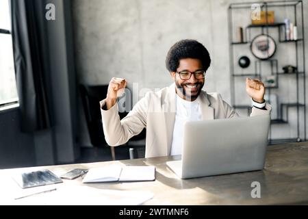 Ein brasilianischer Geschäftsmann wirft seine Hände in die Luft vor seinem Laptop und zeigt einen Moment des Erfolgs an einem modernen Arbeitsplatz, der den Nervenkitzel einer gut erledigten Arbeit symbolisiert Stockfoto