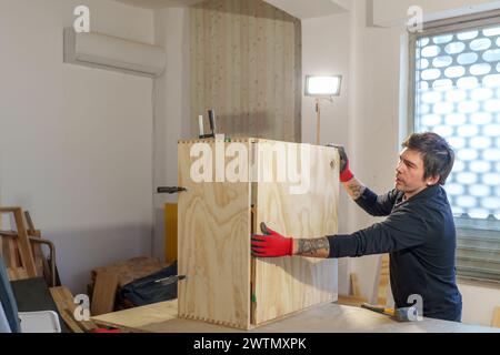Ein hispanischer Zimmermann mittleren Alters, der in seiner Tischlerei steht und ein Möbelstück zusammenbaut Stockfoto
