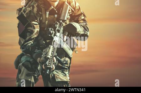 Der Soldat bei der Ausführung von Aufgaben in Tarnung und Schutzhandschuhen, die eine Waffe vor dem Hintergrund des Sonnenuntergangs halten. Kriegsgebiet. Stockfoto