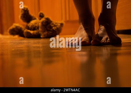 Ein brauner Kinderbär sitzt nachts auf dem Boden eines Hauses in einer Wohnung und neben den Beinen einer Frau, ein Kinderspielzeug Stockfoto