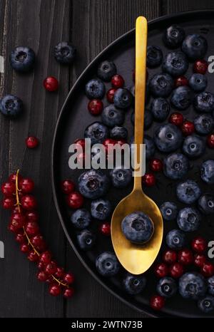 Der goldene Metalllöffel, die Schreibtischoberfläche aus schwarzem Holz und frische natürliche Beeren: Heidelbeeren und rote Johannisbeere auf schwarzem Gericht auf einem Tisch. Stockfoto