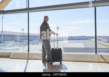 Ein ruhiger männlicher Tourist steht am Flughafen und schaut den Flugzeugflug durch das Fenster. Er hält den Koffer bei Sonnenuntergang Stockfoto