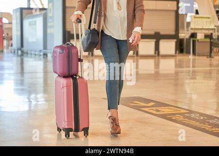 Detail der Beine einer Frau, die mit Koffern mit Gepäck durch das Flughafenterminal geht. Unerkennbare Frau in lässiger Kleidung Stockfoto