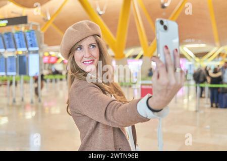 Schöne Frau mittleren Alters, die am Flughafen ein Selfie mit ihrem Smartphone macht. Porträt einer eleganten Frau mit Hut Stockfoto