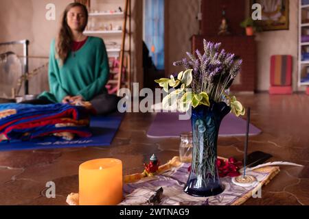 Meditationsaltar mit Lavendelblumen und Räucherstäbchen, mit einer jungen Frau, die im Hintergrund meditiert, außer Fokus. Stockfoto