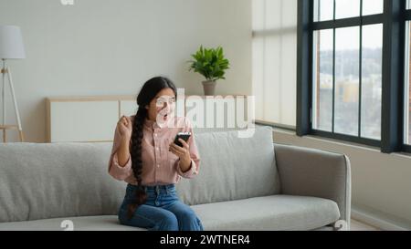 Überglückte Frau Mädchen Frau auf Sofa benutzen Handy aufgeregt Gewinner Kunde erhalten E-Commerce Shopping Angebot gewinnen Bid Jump lustig tanzen auf Stockfoto