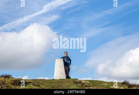 Ein Mann lehnt sich an den Trig Point am Eyam Moor an der William Hill Road, während er im Derbyshire Peak District spaziert und zurückpackt Stockfoto