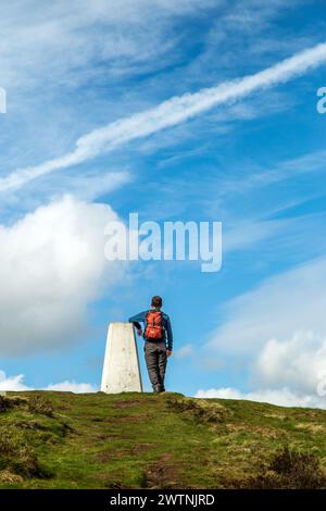 Ein Mann lehnt sich an den Trig Point am Eyam Moor an der William Hill Road, während er im Derbyshire Peak District spaziert und zurückpackt Stockfoto