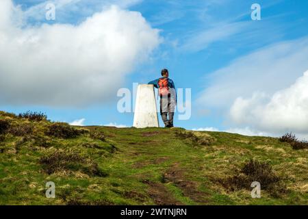 Ein Mann lehnt sich an den Trig Point am Eyam Moor an der William Hill Road, während er im Derbyshire Peak District spaziert und zurückpackt Stockfoto