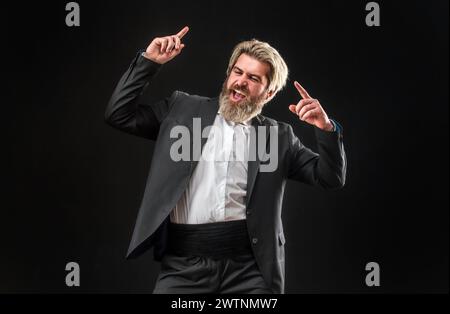 Fröhlicher, junger Geschäftsmann in schwarzem Anzug, der Musik mit Kopfhörern hört und auf grauem Hintergrund tanzt. Stockfoto