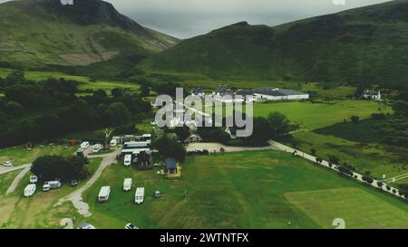 Schottisches Dorf aus der Vogelperspektive: Straße, Hütten, Häuser, Destillerie, Camping im grünen Tal an wolkigen Sommertagen. Malerische Berglandschaft von Arran Island, Schottland. Filmmaterial wie im Kino Stockfoto