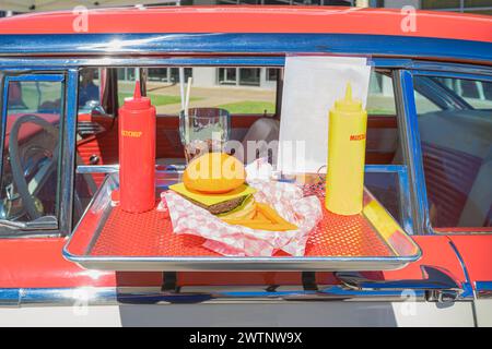 „Classic Drive-in Nostalgie: Ein roter und weißer Oldtimer mit einem Essenstablett am Fenster zeigt ein typisch amerikanisches fest der Cheeseburger Stockfoto