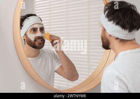 Mann mit Stirnband, der sein Gesicht mit einem Schwamm in der Nähe des Spiegels im Badezimmer wäscht Stockfoto