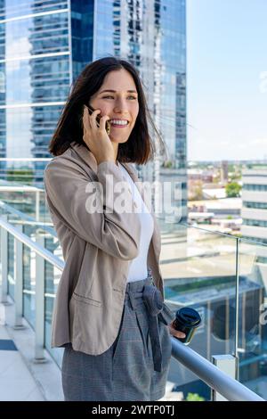 Eine Frau auf ihrem Balkon spricht auf ihrem Handy und hält während ihrer Pause eine Tasse Kaffee in der Hand. Stockfoto