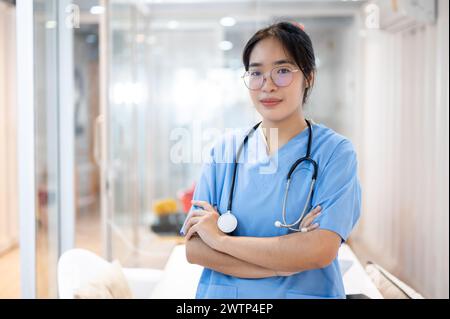 Eine professionelle und selbstbewusste asiatische Ärztin in Peelings, die mit überkreuzten Armen im Krankenhausflur steht. Medizin- und Gesundheitskonzepte Stockfoto