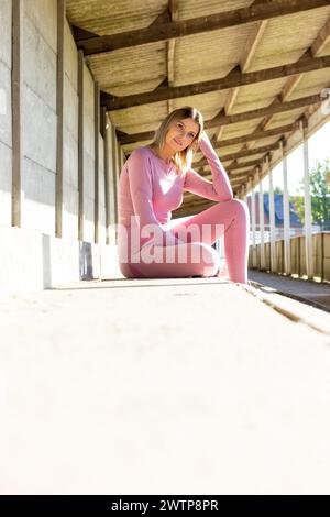 Dieses Foto in einem sonnendurchfluteten Korridor zeigt eine junge Frau, die sich einen Moment in ihrer leuchtenden rosa Aktivkleidung entspannt. Sie sitzt bequem auf dem Betonboden und lehnt sich an eine Säule, die den Rand des Korridors markiert. Die Holzkonstruktion des Dachs erzeugt lineare Schatten, die der Szene Tiefe und Kontrast verleihen. Ihre Haltung ist entspannt, aber balanciert, mit einer Hand, die ihren Kopf stützt, was auf eine Pause von körperlicher Aktivität oder Kontemplation hindeutet. Ihr Blick auf die Kamera ist freundlich und einladend. Das warme Sonnenlicht taucht den Flur auf und erzeugt einen hochmodernen Lichteffekt, der ihn hervorhebt Stockfoto