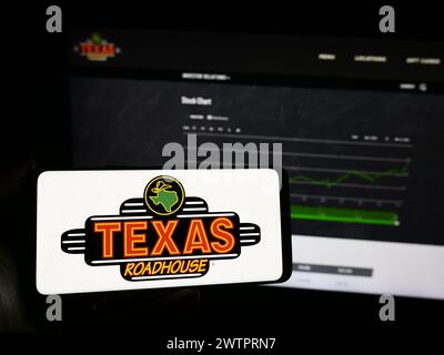 Person mit Mobiltelefon mit Logo des amerikanischen Steakhouse-Restaurantunternehmens Texas Roadhouse Inc. Vor der Webseite. Schwerpunkt auf dem Display des Telefons. Stockfoto