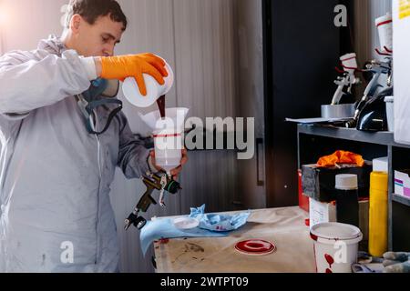 Lackierer in Schutzkleidung gießt Mischfarbe aus einer Dose in einen Kunststoffmischbehälter an einer Kfz-Tankstelle. Stockfoto