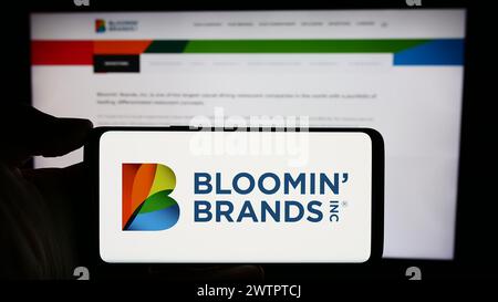 Person, die ein Mobiltelefon mit dem Logo der US-Restaurantholding Bloomin' Brands Inc. Vor der Webseite des Unternehmens hält. Schwerpunkt auf dem Display des Telefons. Stockfoto