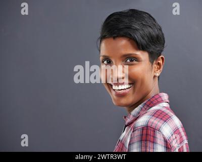 Glückliche, Portrait und indische Person mit einem Lächeln mit grauem Hintergrund in einem Büro einer Kreativagentur. Hipster, Selbstvertrauen und nicht binäre Mitarbeiter mit Moderne Stockfoto