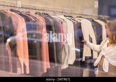 Frau kauft Kleidung in einem Geschäft, viele verschiedene Stile und Farben zur Auswahl. Verschwommene Ansicht im Speicherfenster, selektiver Fokus Stockfoto