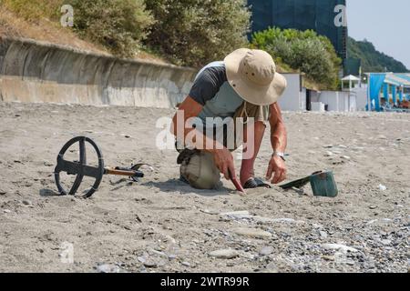 Ein Mann mit Shorts und Hut, mit Metalldetektor, sucht tagsüber im Sommer an einem Sandstrand nach Gold und Münzen. Stockfoto