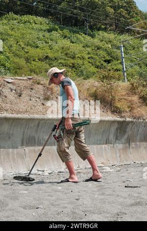 Ein Mann mit Shorts und Hut, mit Metalldetektor, sucht tagsüber im Sommer an einem Sandstrand nach Gold und Münzen. Stockfoto