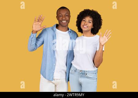 Glückliches afroamerikanisches Paar, das die Hände mit hellem Lächeln zur Kamera winkt, lässig gekleidet Stockfoto