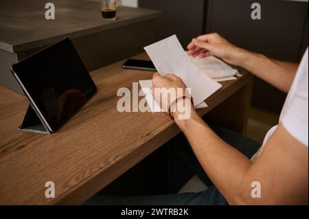 Nahaufnahme eines Mannes, der in seinem Heimbüro sitzt und Rechnungen in Händen hält und online über ein digitales Tablet bezahlt. E-Banking, Internet, mobiles Banking A Stockfoto