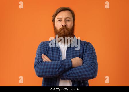 Rothaariger bärtiger Mann, der die Arme auf der Brust vor orangefarbenem Hintergrund kreuzt Stockfoto