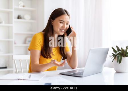 Selbstbewusste junge asiatische Freelancerin, die auf Handy in der Nähe eines offenen Laptops spricht Stockfoto