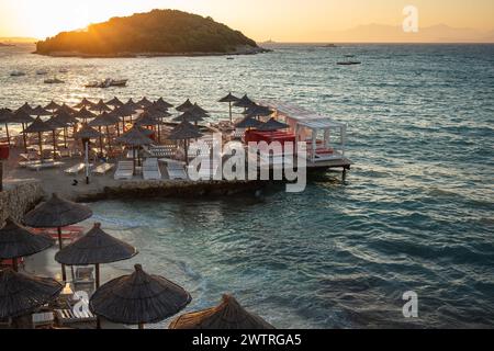 Strandlandschaft von Ksamil Beach mit seiner Insel bei Sonnenuntergang. Sommerabend mit Ionischem Meer und Sonnenliegen mit Sonnenschirmen an der albanischen Riviera. Stockfoto
