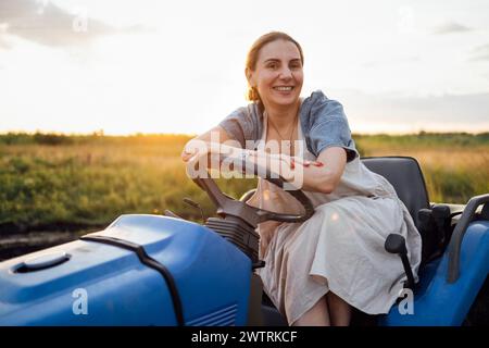 Ein junger, lächelnder Bauer sitzt am Lenkrad eines Traktors. Eine lustige charmante Frau in lässiger Kleidung bewirtschaftet ein landwirtschaftliches Feld. Erntezeit. Sonnenuntergang Stockfoto