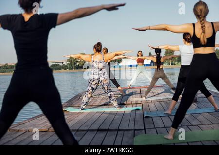 Yogakurs am Vormittag im Freien mit Lehrer in der Nähe des Flusses in der Stadt. Junge Frauen stehen mit ausgestreckten Armen auf Matten und machen Übungen. Gruppe von Mult Stockfoto