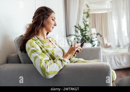 Eine charmante junge Frau hält ein Smartphone und sitzt auf der Couch im Wohnzimmer. Eine attraktive Brünette liest die Nachrichten und kauft usi Stockfoto