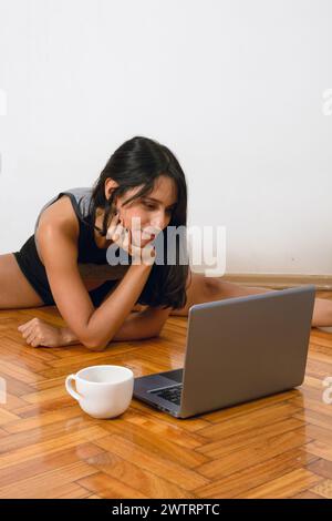 Vertikales Bild der jungen hispanischen Latina Brünette Frau mit schwarzen Haaren und Sportbekleidung, sitzend auf dem Boden, glücklich, Yoga-Kurse mit Laptop zu Hause Stockfoto