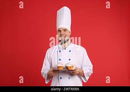 Glücklicher professioneller Konditor in Uniform mit köstlichen Cupcakes auf rotem Hintergrund Stockfoto