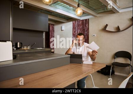 Gestresster und besorgter Kerl, der auf dem Handy spricht, streitet, während er unzufrieden war, indem er Rechnungen bezahlt, Papierkram macht, am Küchentisch zu Hause sitzt Stockfoto