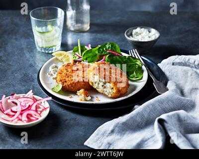 Knuspriger panierter Fisch mit einer Seite von frischem Spinatsalat und in Scheiben geschnittenen eingelegten Zwiebeln auf einem dunklen, eleganten Tisch. Stockfoto