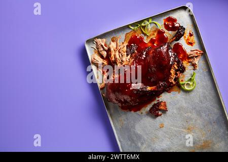 Herzhaftes Pulled Schweinefleisch mit Grillsoße auf einem Metalltablett vor violettem Hintergrund Stockfoto